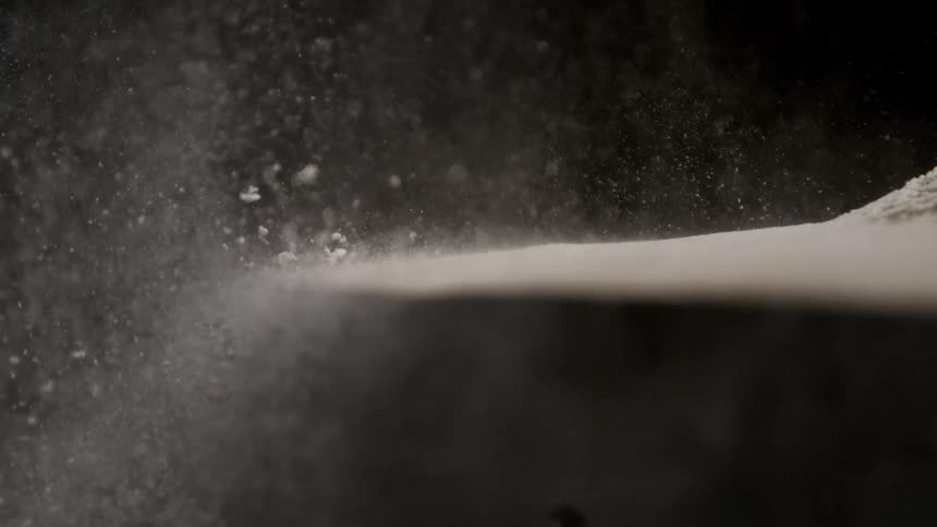 In slow motion a drumstick strikes a powder covered cymbal causing a dramatic explosion of white dust against a black background