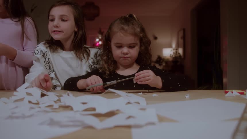 Three young girls are standing near a wooden table and one girl uses a pair of shears to cut folded white paper while a pile of cut paper pieces rests on the table surface