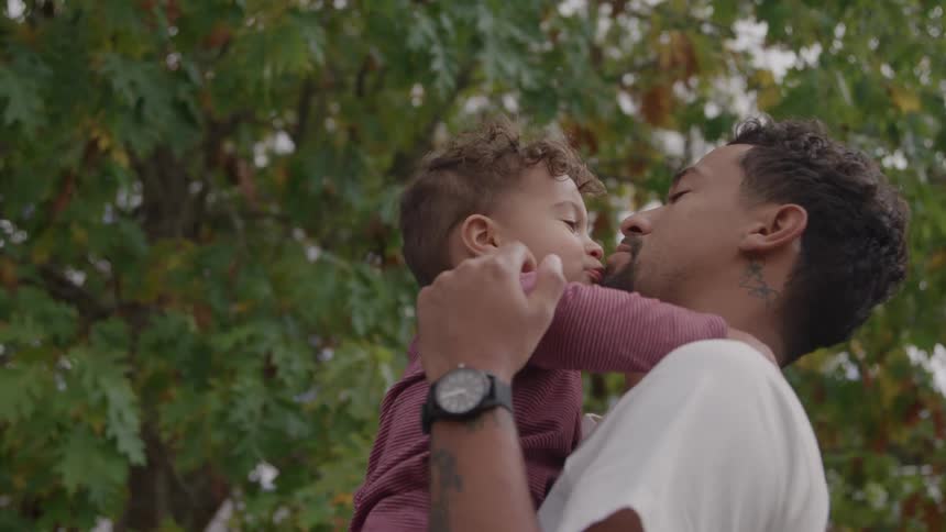 A man holding a small child in his arms with the child facing him and hugging his neck then the man smiles and the child leans in for a kiss against a backdrop of green tree leaves