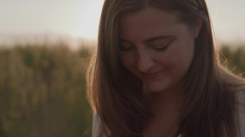 A young woman with long hair smiles gently as she looks down backlit by the warm glow of the setting sun