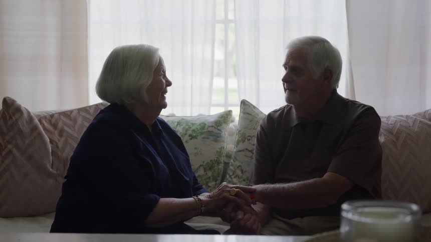 An elderly couple with gray hair sitting close together on a couch facing each other holding clasped hands and smiling while looking at each other backlit by a bright window covered with sheer white curtains