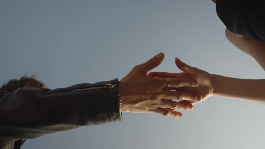 Two human forelimbs extend toward one another against a clear sky background resulting in the hand of a person wearing a dark sleeve and a ring briefly connecting with the lower arm of another person