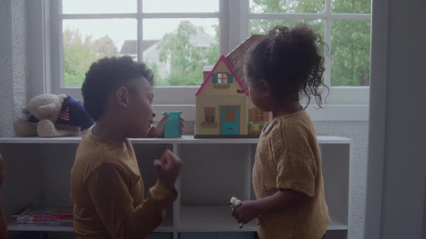 Two kids play with a dollhouse beside a window at midday