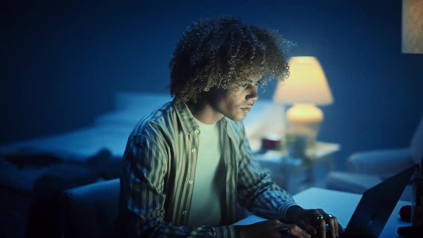 A young man lit by a warm lamp works intently on a laptop in a dim blue bedroom