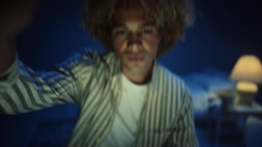 Person looks directly forward in a room lit by soft blue light with a lamp glowing in the background