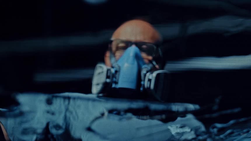 A bald man wearing safety glasses and a blue respirator mask covers a translucent surface with a clear coating in a close view wearing a blue shirt in a dark industrial setting