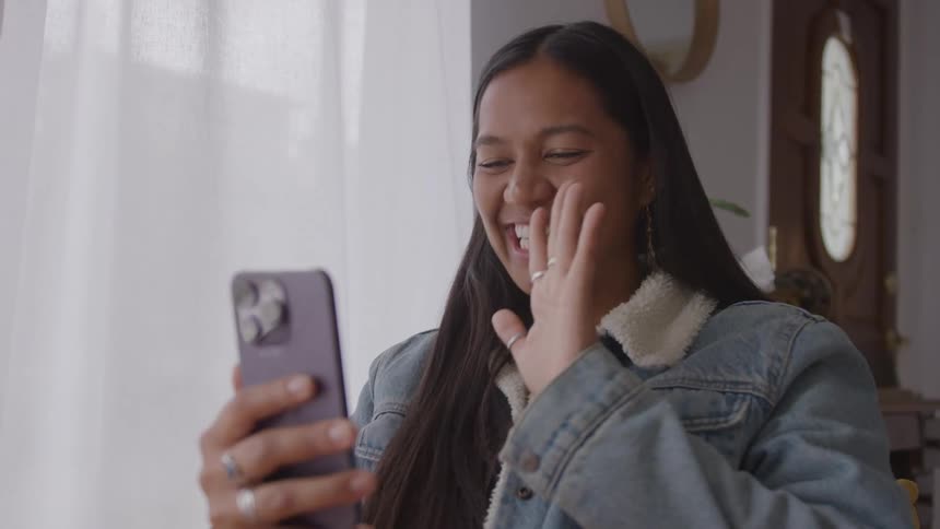 The video shows a smiling young woman with long dark hair engaging in a video call on her smartphoneshe enthusiastically waves her hand at the camera on her phone wearing a denim jacket with a sherpa collar in an indoor setting