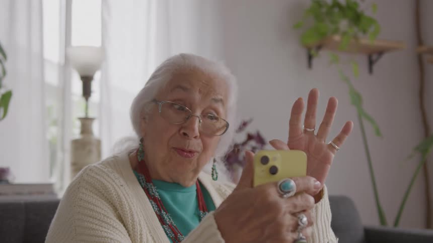 The video features a closeup of an elderly woman with long grey hair and glasses holding a yellow smartphone for a video callshe is smiling and enthusiastically waving her hand toward the phone wearing a white cardigan and several pieces