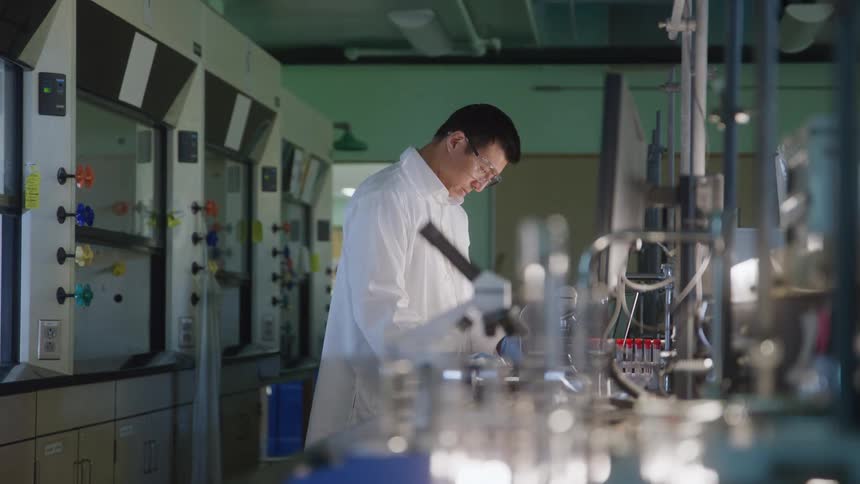 In a modern laboratory a male scientist in a white coat and safety glasses is focused on his work while a female colleague is in the foreground