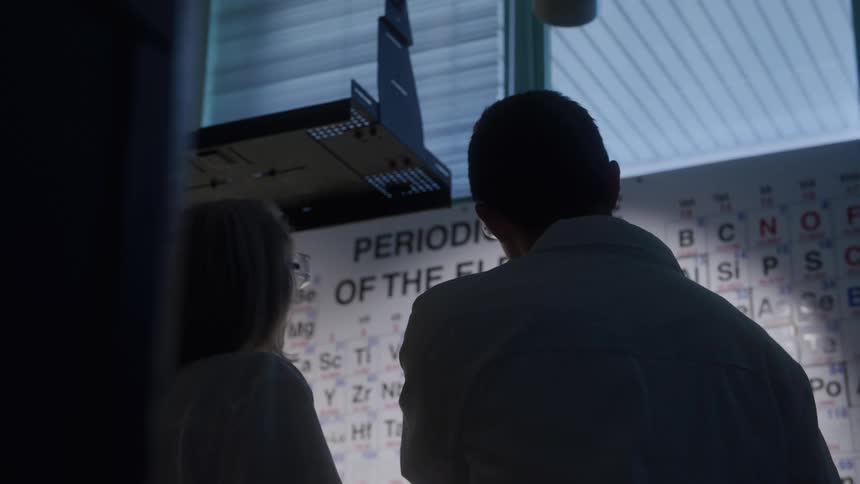 The video shows the silhouettes of two scientists facing away from the camera looking at a large wallmounted periodic table of the elementsone scientist on the left an older woman points towards a part of the chart while speaking to th