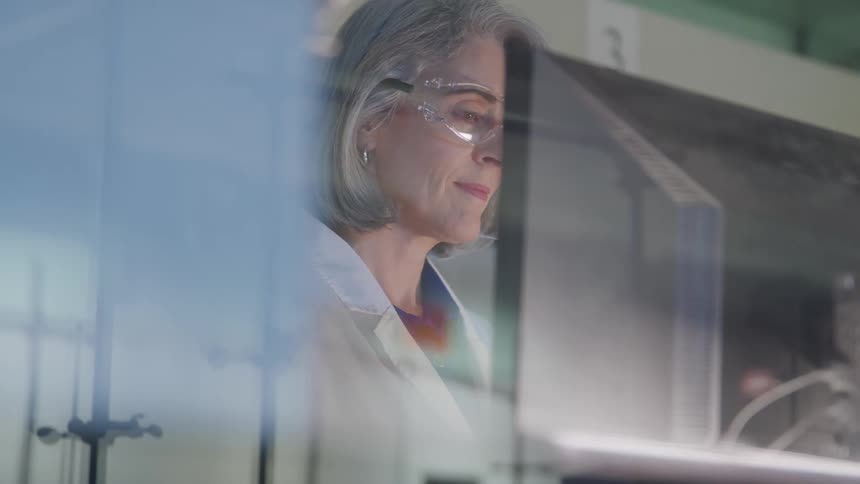 The video features an older female scientist in a lab coat and safety glasses working at a computershe is concentrating on the monitor which is partially obscured in the foreground creating a reflection on the glass barrier in front of her
