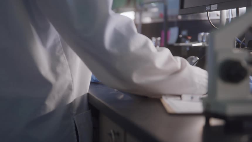 The video is a closeup of a person in a lab coat and blue nitrile gloves typing on a computer keyboardthe work is taking place in a lab with a microscope and other equipment visible on the countertop next to the keyboard