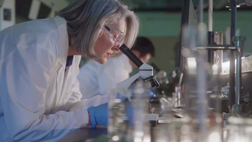 In a science lab a male researcher holds a test tube up to the light for examination while his female colleague walks into the foreground