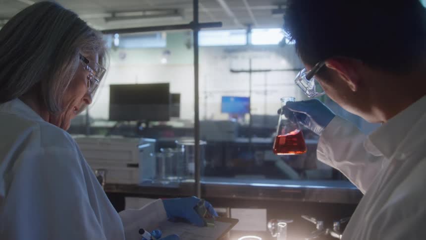 Man and woman conducting laboratory analysis with red liquid in a flask while recording data on a clipboard