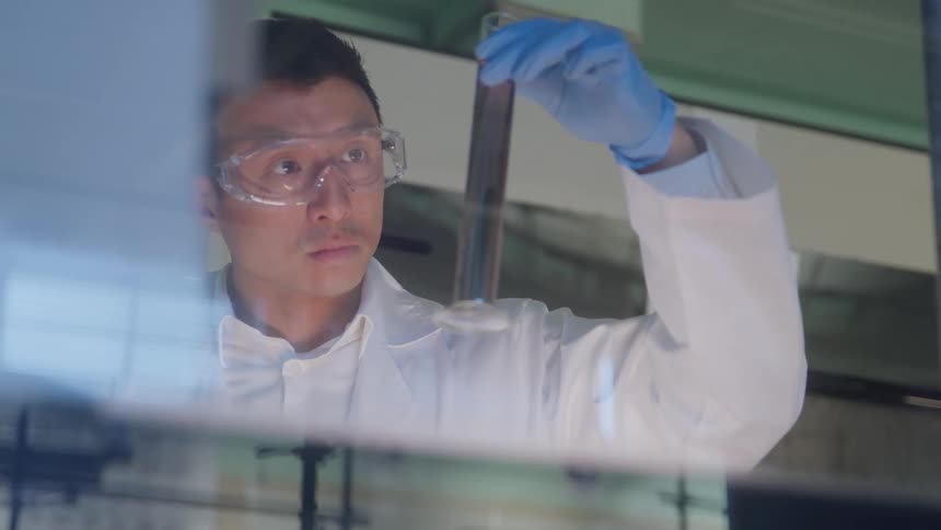 The video shows a male scientist in a lab coat and safety goggles examining a substance in a test tubehe is holding the test tube which appears to contain a dark reddishpurple liquid up to the light for close inspection with a blue