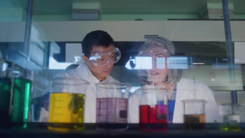 Man and woman observing colorful liquids in beakers lined up on a laboratory bench with scientific equipment in the background