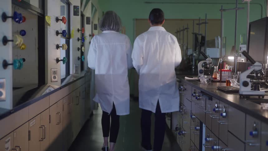 The video shows two scientists a man and a woman walking away from the camera down an aisle in a laboratoryboth are wearing white lab coats and are moving past a long row of lab benches with drawers and various scientific equipment