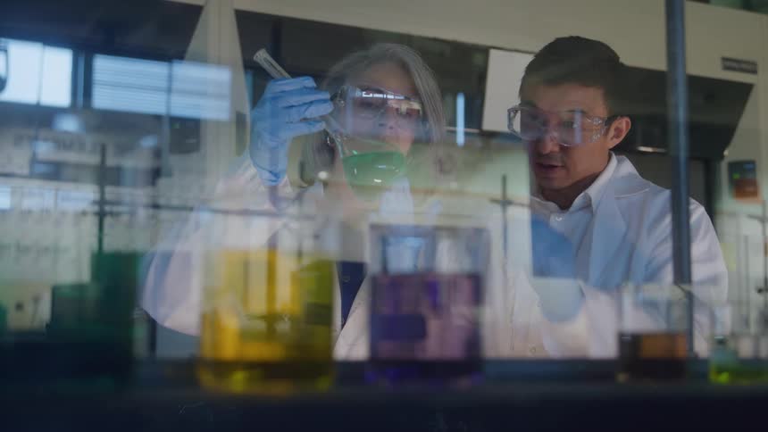 A young male and female scientist examine samples in a beaker both wearing white lab coats and goggles as they engage in a focused discussion during the process