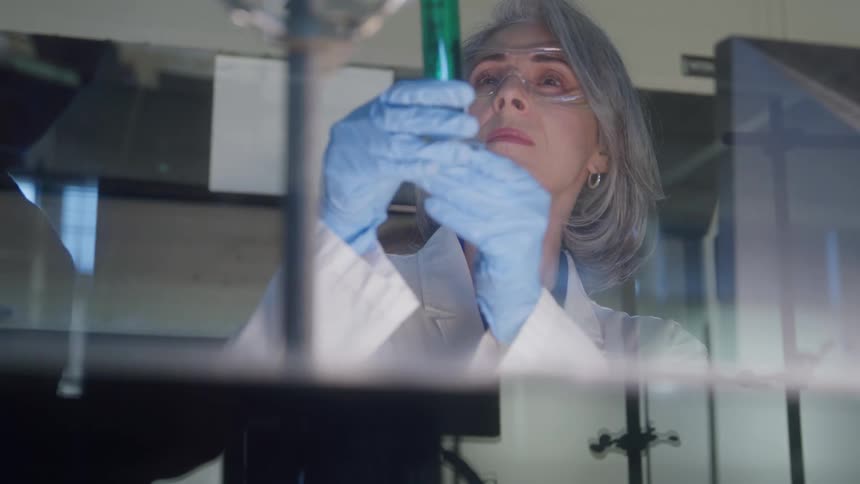 A female scientist in safety goggles and a lab coat carefully examines a test tube filled with a green liquid