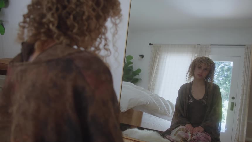 A woman with long curly blonde hair wears a brown jacket and sits on the floor looking at her reflection in a large mirror inside a bright sunlit bedroom