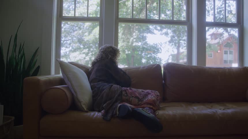 The video shows a woman seated indoors on a light brown leather couch wrapped in a blanket looking out a large window the lighting is dim inside highlighting the bright green trees and light from the windows creating a mood of quiet contemplati