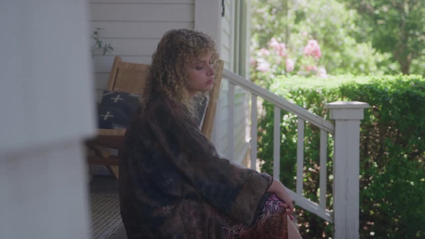 A woman with long curly blonde hair wears a patterned kimono and sits on the wooden porch of a white house with a green hedge and pink flowers visible in the sunlit background