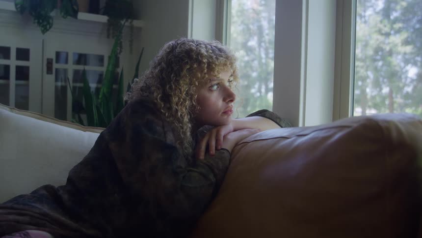A young woman looking through a window contemplatively while sitting in a living room during daytime