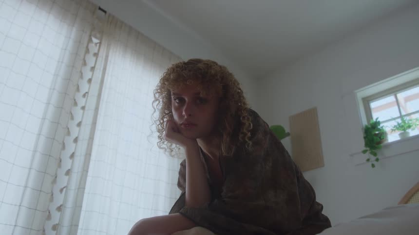 The video features a young woman with curly reddishblonde hair sitting indoors near a window with sheer curtainsshe is resting her chin on her hand looking directly at the camera with a solemn or introspective expression