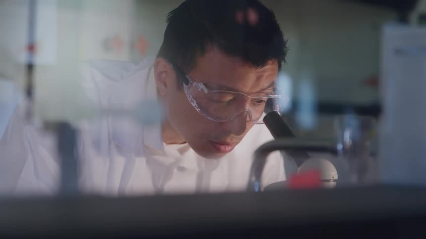 A young male lab scientist wearing goggles and a lab coat peers into a microscope with focused attention
