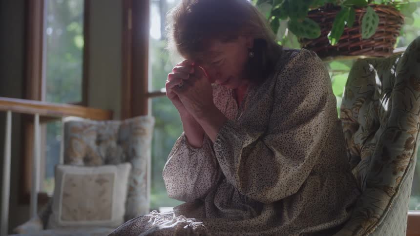 Woman sitting on a chair with her hands clasped and head bowed expressing sadness