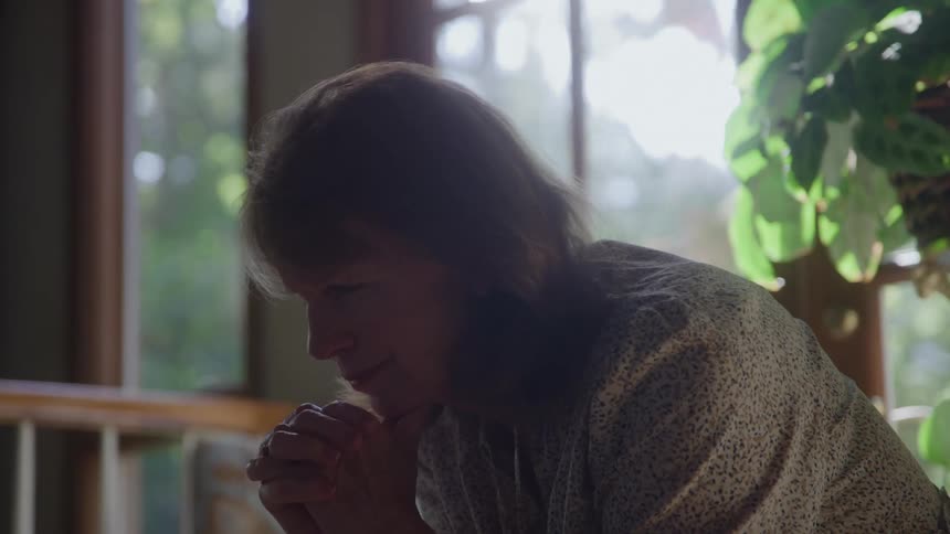 A close profile view of an older woman with her eyes closed and hands clasped shows her praying or meditating in front of a brightly sunlit window with green foliage visible outside