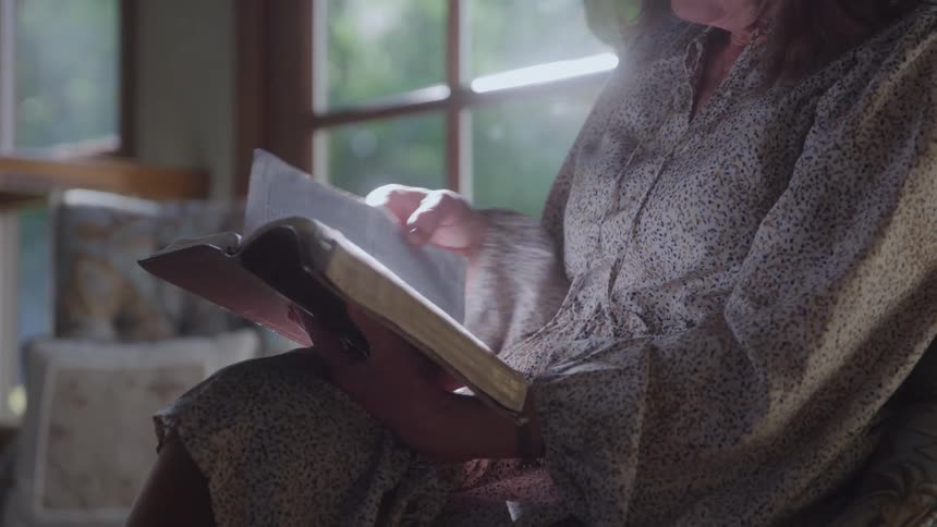 Person sitting by a window reading a thick book in an indoor setting lit by natural light