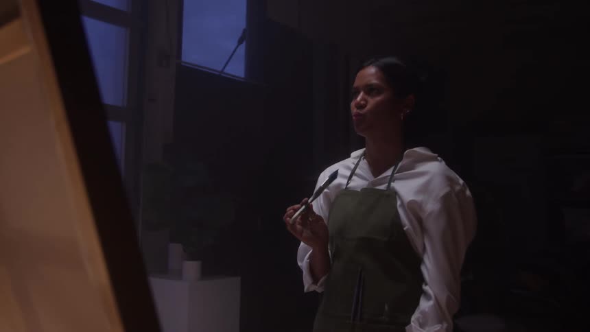 A young woman stands in a studio holding a paintbrush gazing thoughtfully at her canvas as she contemplates her artwork