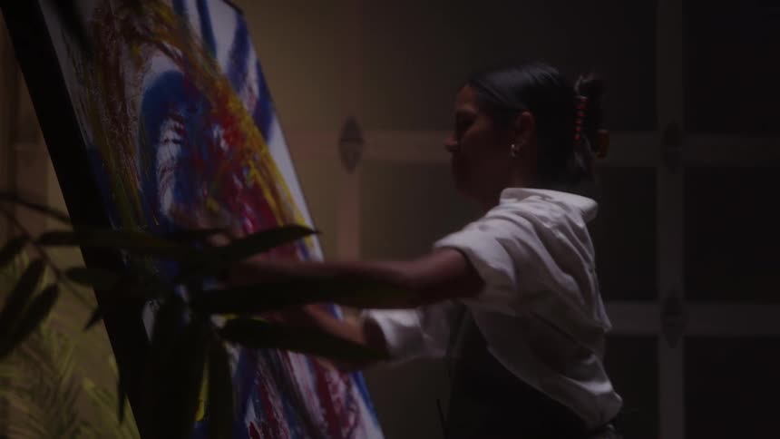 A rear profile close view shows a woman artist in a white shirt and black apron painting dynamic blue yellow and red strokes onto a tall canvas in a dark studio