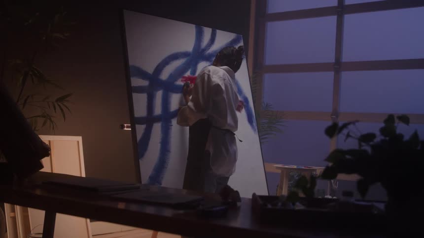 A young female artist paints on a canvas holding a brush in one hand and a paint can in the other applying bold strokes of blue paint with focused intent