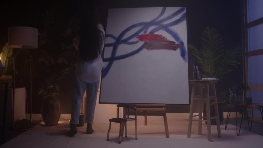In a dimly lit studio a female artist walks up to a large abstract canvas and puts on her apron to begin painting