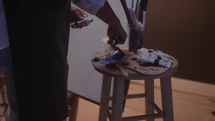 A close view shows an artist in a dark apron squeezing paint from a tube onto a wooden palette that rests on a stool surrounded by paint tubes and brushes in jars