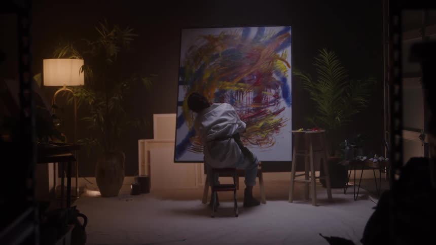 Woman seated in front of the canvas painting colorful shapes in an artistic setting with warm lighting and surrounding plants