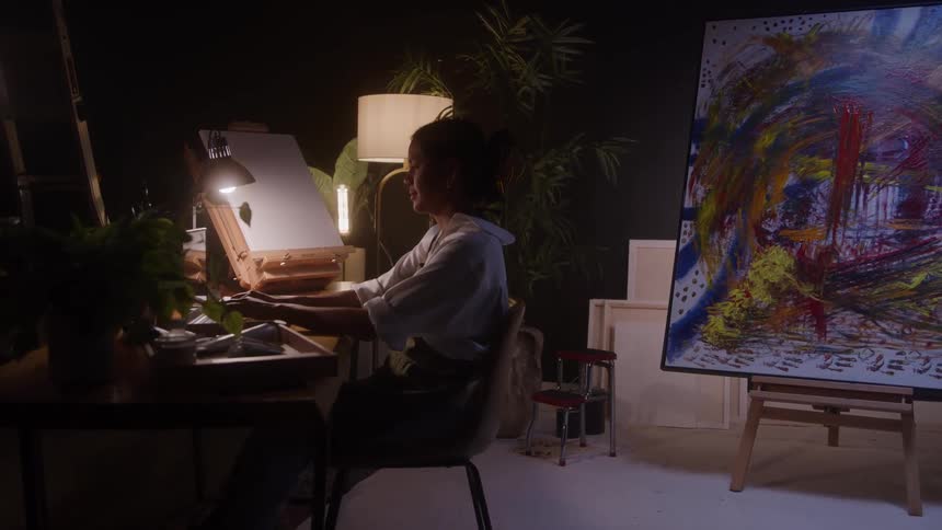 The video shows an artist working in a dimly lit studio focused on a desk with a canvas and a small lampin the foreground a large abstract painting with bold brushstrokes of red yellow and black on a white canvas is prominently displayed on an
