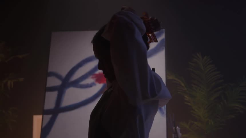 Woman ties her hair in front of a partially painted canvas under soft lighting
