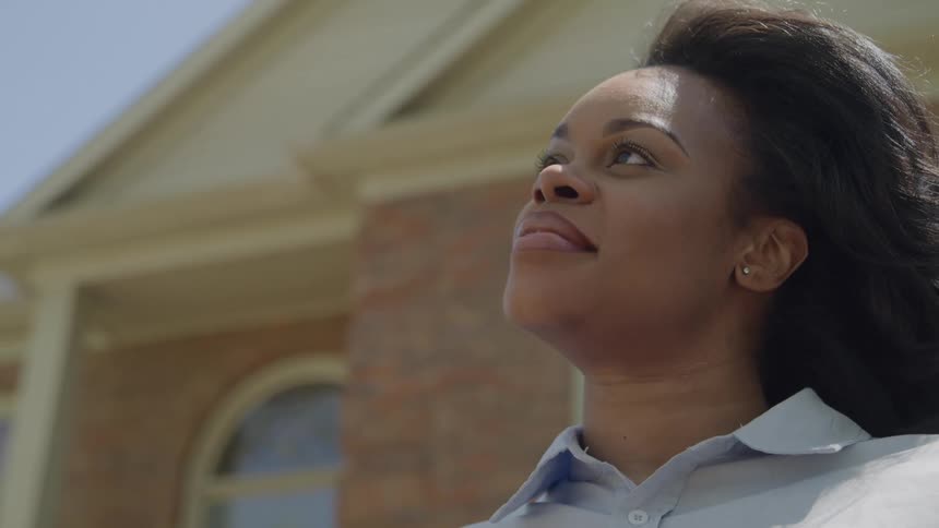 A young black woman with long black hair stands outdoors in front of a building bathed in sunlight