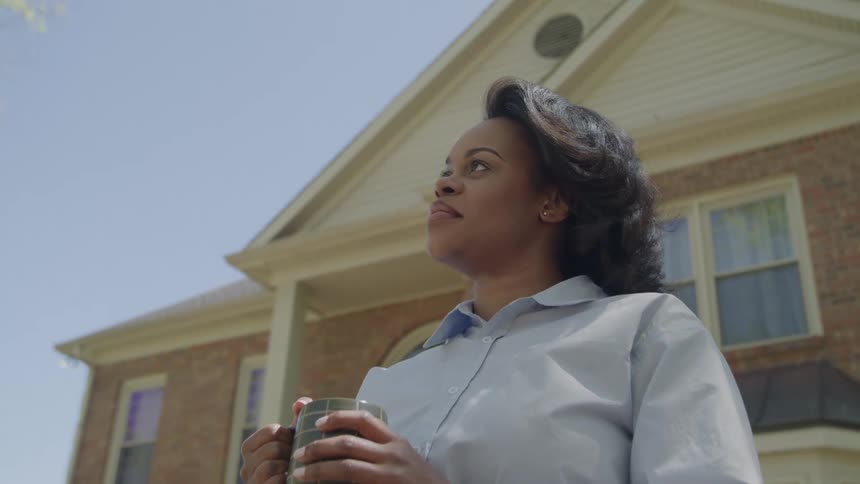The video shows an african american woman standing outdoors in front of a brick house holding a mugshe is looking up and away from the camera wearing a light blue or gray collared shirt with a contemplative or hopeful expression