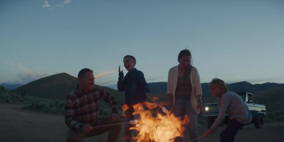 The video shows a family of fourtwo adults and two childrenroasting marshmallows over a campfire at duskthey are smiling and gathered around the brightly burning flames outdoors with a mountain range visible in the background under a pale bl