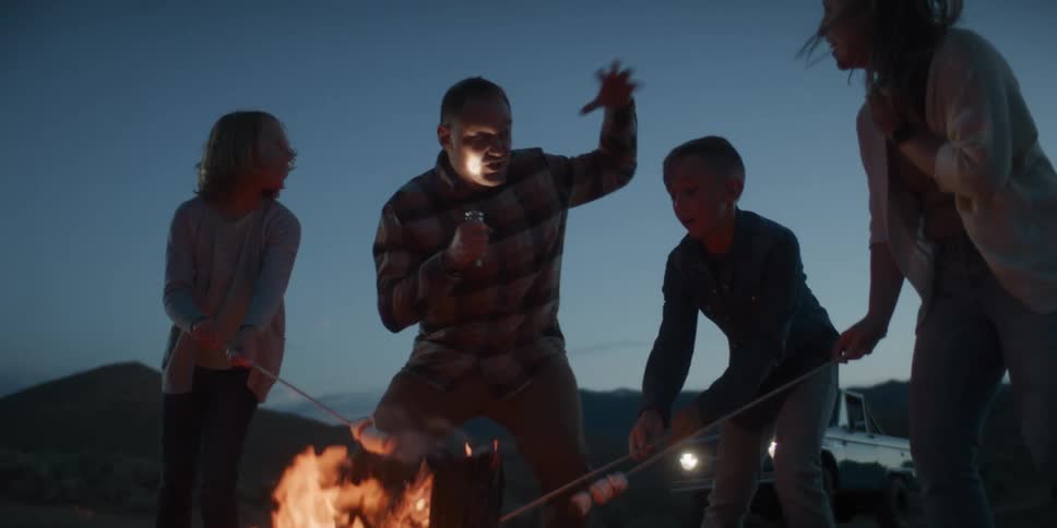 Family gathered around a campfire at dusk sharing laughter and marshmallows in a natural setting with a vehicle in the background