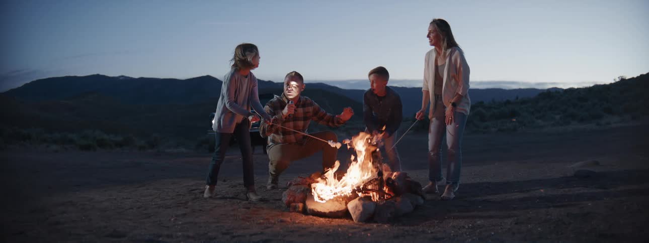 A family enjoys roasting marshmallows over a campfire at dusk with the father playfully telling a scary story