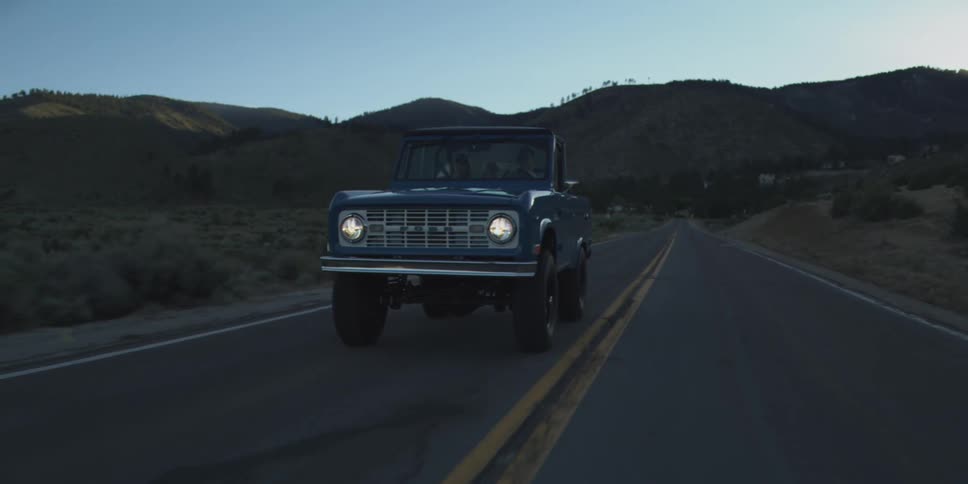 A dark blue vintage offroad vehicle driving down a paved two lane road at dusk with its headlights on surrounded by dark hills and dry brush
