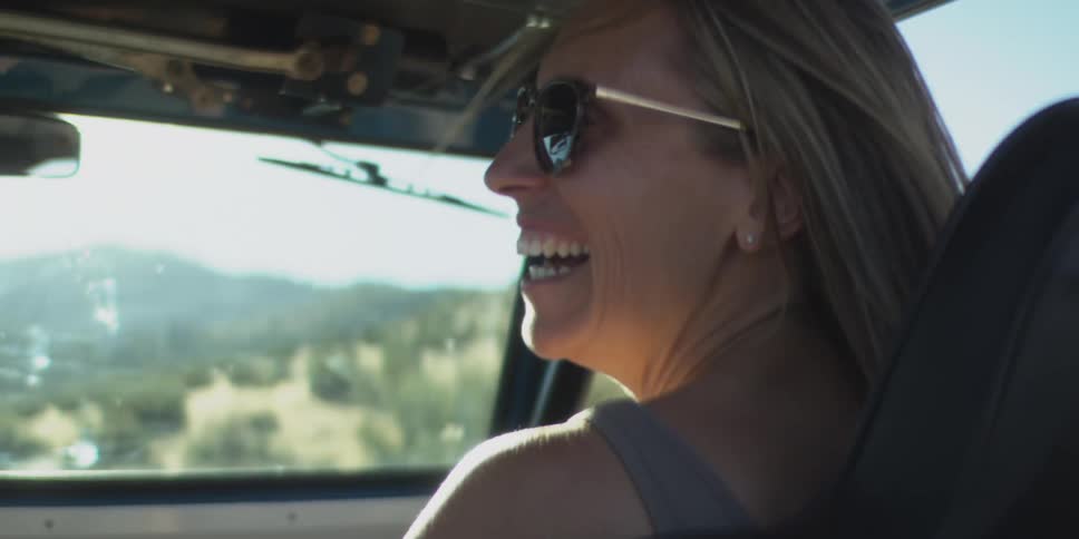 The video is a closeup of a woman smiling cheerfully while driving an opentop vehicle on a sunny open roadshe is wearing a tank top and sunglasses and her long hair is pulled back as she enjoys the drive through the scenic rolling hills
