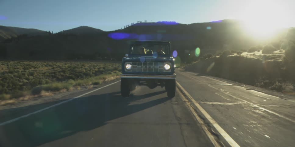 The video shows an old dark blue suv or offroad vehicle driving down a paved road directly towards the camera the scene is set in a mountainous scrub brush landscape on a sunny day with a significant sun flare visible on the right