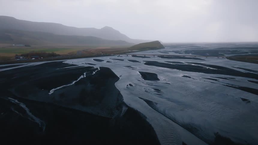 Aerial view of winding river channels flowing through dark volcanic sand under overcast sky