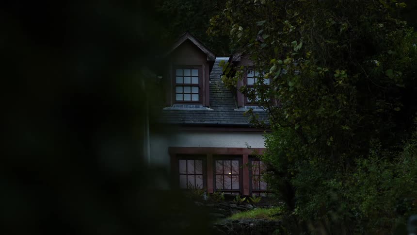 A view through dark foreground trees slowly reveals a secluded twostory house with dormer windows and lit lower windows sitting on a stone base against a backdrop of dense foliage